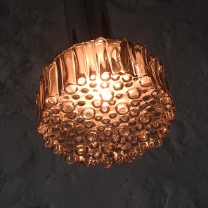 Puede incluir: Una lámpara colgante de cristal color ámbar de estilo vintage. La luz tiene una superficie texturizada con forma de burbujas y un soporte metálico cilíndrico. La luz se ilumina desde el interior, emitiendo un brillo cálido.