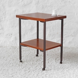 May include: A two-tiered side table with a dark brown wooden top and shelf, supported by a dark metal frame. The table has small wheels on the bottom of each leg. A white mug with a black design sits on the top shelf.