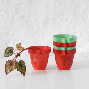 Könnte beinhalten: Eine Sammlung roter und grüner gerippter Pflanztöpfe aus Kunststoff. Ein Topf steht einzeln, die anderen sind gestapelt. Eine kleine Pflanze mit grün-rot gefleckten Blättern befindet sich neben den Töpfen. Die Töpfe sind für den Innen- oder Außenbereich geeignet.