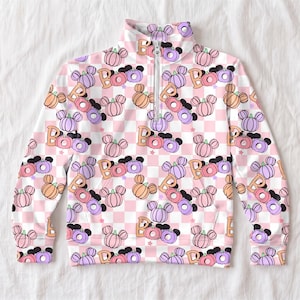 May include: A long-sleeve quarter-zip pullover featuring a repeating pattern of pastel pink, orange, and purple pumpkins with mouse ears, interspersed with the word Boo in orange, all set against a light pink and white checkered background.