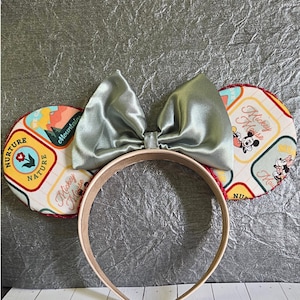 May include: A handmade headband featuring Mickey Mouse ears. The ears are round with a colorful pattern and the text "Nurture Nature" and "Mickey Mouse". A large, light green satin bow sits atop the headband. The headband is tan with a red trim.