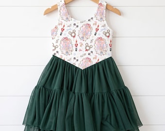 May include: A sleeveless dress featuring a white bodice with a repeating print of nutcrackers, castles, and mouse-ear wreaths in red, pink, and gold. The waistline is V-shaped, connecting to a layered, dark green tulle skirt that falls to the knee.