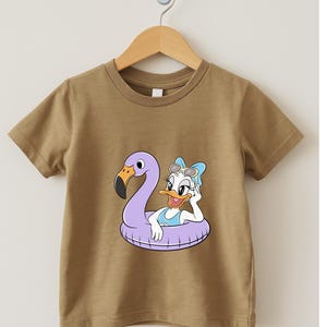 May include: A tan short-sleeved t-shirt featuring a cartoon graphic of Daisy Duck in a flamingo-shaped inflatable ring. Daisy Duck is wearing sunglasses and a blue swimsuit. The t-shirt is hanging on a wooden hanger.