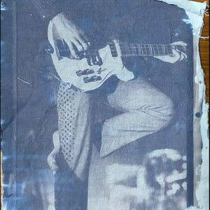 May include: A black and white photo of a person playing a bass guitar. The photo is printed on a piece of fabric.