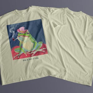 May include: A light green frog wearing a pink hat and smoking a pipe. The frog is sitting on a red and pink surface. The image is on a light beige t-shirt with the text "this is not a frog".