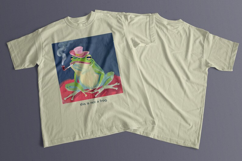 May include: A light green frog wearing a pink hat and smoking a pipe. The frog is sitting on a red and pink surface. The image is on a light beige t-shirt with the text "this is not a frog".