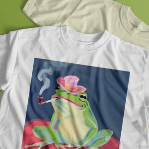 May include: White t-shirt with a graphic of a green frog wearing a pink hat and smoking a pipe. The frog is sitting on a red and pink background.