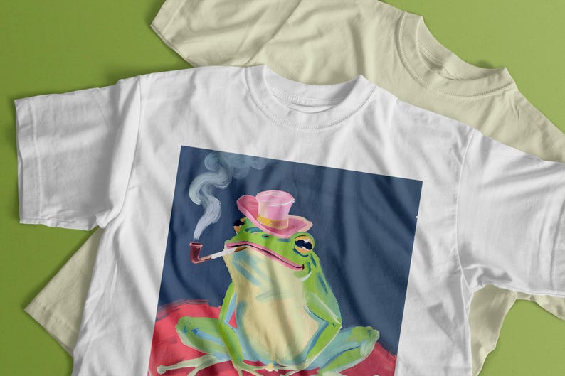 May include: White t-shirt with a graphic of a green frog wearing a pink hat and smoking a pipe. The frog is sitting on a red and pink background.