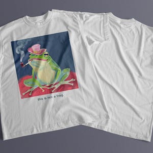 May include: White t-shirt with a graphic of a green frog wearing a pink hat and smoking a pipe. The text "this is not a frog." is printed below the frog.