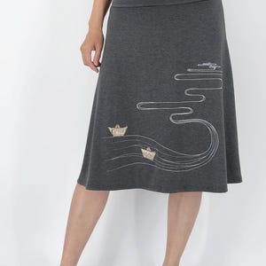 May include: A gray skirt with a flowing design of white lines and two small boat illustrations. The skirt is knee-length and has a wide waistband. The model is wearing white sneakers.