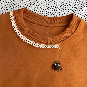 Op de afbeelding: Een roestkleurig sweatshirt met een geribbelde ronde halslijn versierd met een delicate crèmekleurige kanten rand. Een kleine zwarte knoop in de vorm van een spin met witte ogen is aan de stof bevestigd, verbonden door een dunne geborduurde lijn.