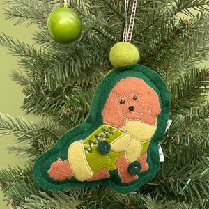 May include: A felt Christmas ornament shaped like a dog, with brown fur and wearing a green sweater. The ornament is trimmed in dark green and has a small green pom-pom at the top. It hangs from a ribbon on a Christmas tree.