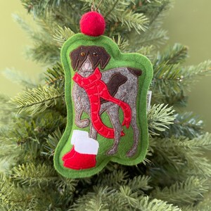 May include: A felt Christmas ornament featuring a dog wearing a red scarf and boots. The ornament is green with a red pom-pom at the top. The dog is gray and brown, with a red scarf and boots.