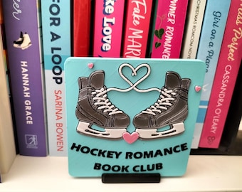 May include: A teal-colored sign with the words "HOCKEY ROMANCE BOOK CLUB" and an illustration of two gray ice skates connected by a heart-shaped design. Pink hearts are also present.