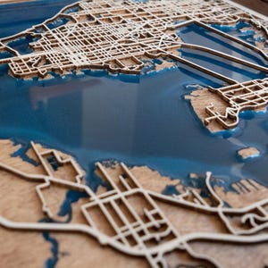 Op de afbeelding: Een gedetailleerde houten kaart met een stadsplattegrond, met ingewikkelde straatpatronen en kustlijnen. De watergebieden zijn gevuld met een doorschijnende blauwe hars, waardoor een 3D-effect ontstaat. Een uniek kunstwerk.