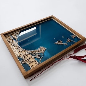 Op de afbeelding: Houten lijst met kaart van Port Clinton, Ohio, met een blauw hars water effect. De kaart is lasergesneden met gedetailleerde weergave van de stad en de omliggende eilanden. De lijst is van natuurlijk hout. Een uniek stukje wandkunst.