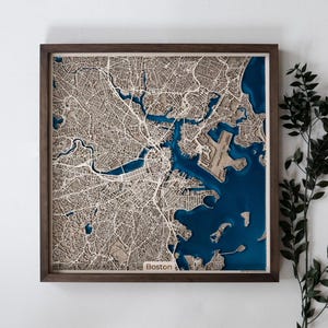 Op de afbeelding: Een driedimensionale houten kaart van Boston, Massachusetts, in een frame. De kaart toont gedetailleerde lasergesneden straten en waterwegen, met diepblauwe hars die het water voorstelt. De lijst is donkerbruin. Het woord "Boston" is zichtbaar.