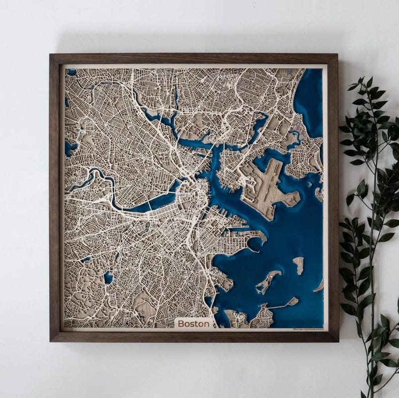 Op de afbeelding: Een driedimensionale houten kaart van Boston, Massachusetts, in een frame. De kaart toont gedetailleerde lasergesneden straten en waterwegen, met diepblauwe hars die het water voorstelt. De lijst is donkerbruin. Het woord "Boston" is zichtbaar.