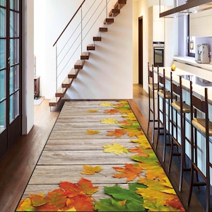 Puede incluir: Una alfombra rectangular para interiores con un diseño de tablones de madera y hojas de otoño dispersas en tonos rojos, amarillos y verdes. La alfombra está colocada en un pasillo, con una escalera y una cocina visibles al fondo.
