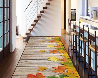 Puede incluir: Una alfombra rectangular para interiores con un diseño de tablones de madera y hojas de otoño dispersas en tonos rojos, amarillos y verdes. La alfombra está colocada en un pasillo, con una escalera y una cocina visibles al fondo.
