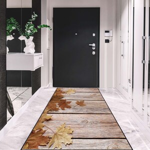 Puede incluir: Una alfombra rectangular con diseño de vetas de madera y hojas de otoño dispersas. La alfombra presenta una impresión realista de tablones de madera desgastados y hojas de arce marrones.