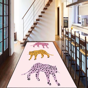 May include: A pink runner rug featuring three leopard illustrations in pink, yellow, and purple with black spots. The rug is rectangular and placed in a hallway with a wooden staircase and kitchen visible in the background.