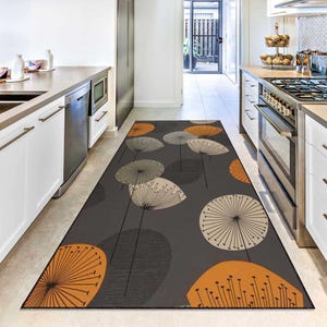 Peut inclure: Tapis de cuisine rectangulaire avec un fond gris foncé et un motif de têtes de graines de pissenlit stylisées en orange, crème et gris. Le tapis est placé dans une cuisine avec des armoires blanches et des appareils en acier inoxydable.