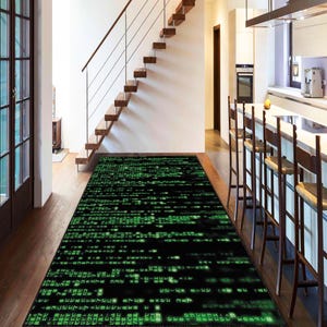 Könnte beinhalten: Ein rechteckiger schwarzer Teppich mit einem grünen digitalen Code-Muster. Der Teppich liegt auf einem Holzboden in einem Flur, mit einer Treppe und einer Küche im Hintergrund. Das Muster des Teppichs ähnelt Computercode.