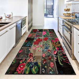Op de afbeelding: Een rechthoekig patchwork vloerkleed met bloempatronen in een keuken. Het kleed heeft bruine, beige en zwarte vlakken, versierd met rode, roze, blauwe en groene bloemmotieven. Het kleed ligt op een lichtgekleurde vloer.