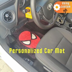Könnte beinhalten: Schwarze Auto-Fußmatte mit einem roten Spider-Man-Masken-Design und weißen Augenformen. Die Matte befindet sich in einem Autoinnenraum, mit Lenkrad und Sitz sichtbar. Der Text auf der Matte lautet "Personalized Car Mat".