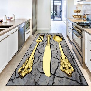 May include: A rectangular kitchen rug with a design of large gold-coloured cutlery: a fork, knife, and spoon. The utensils are set against a grey, cracked earth background, creating a decorative floor covering.