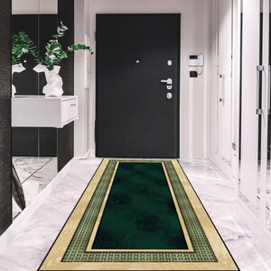 May include: A rectangular hallway rug featuring a dark green central section, bordered by a beige frame and a patterned green and beige trim. The rug is positioned in front of a black door, on a white marble floor.
