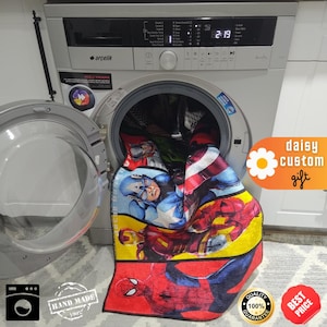 May include: A colorful superhero-themed blanket featuring Captain America, Iron Man, and Spider-Man. The blanket is in a washing machine with the door open. The image also includes the text "daisy custom gift".
