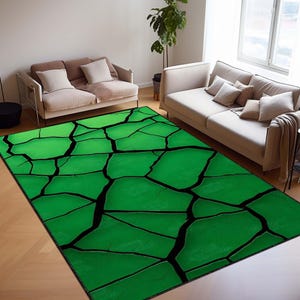 May include: A rectangular green area rug with a cracked, stone-like pattern in black. The rug is placed on a hardwood floor in a living room setting, with two sofas and a window visible in the background. The rug's design gives a modern, abstract look.