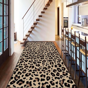 Puede incluir: Una alfombra rectangular con estampado de leopardo. La alfombra tiene un fondo beige con manchas negras y marrones. Se coloca en un pasillo con suelo de madera y una escalera.