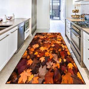 Pode incluir: Um tapete de cozinha retangular com um design de folhas de outono. O tapete apresenta uma mistura de folhas laranja, amarelas, castanhas e vermelhas, criando uma estética acolhedora. O tapete está colocado numa cozinha.