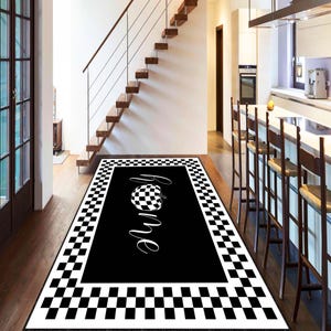 Puede incluir: Alfombra rectangular negra con borde a cuadros y centro negro. El centro presenta la palabra "home" en fuente cursiva, con un diseño de calabaza a cuadros encima de la palabra. La alfombra está colocada sobre un suelo de madera.