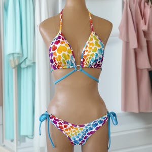 May include: A colorful bikini set on a mannequin, featuring a halter top and tie-side bottoms. The bikini is patterned with rainbow-colored hearts on a white background, with blue accents. The mannequin is displayed in a boutique setting with clothing racks in the background.