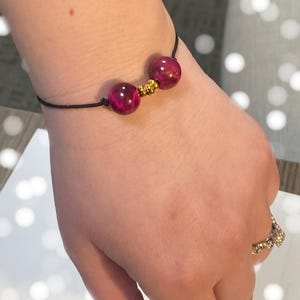 May include: A simple bracelet with two dark pink beads and gold spacers on a black cord.