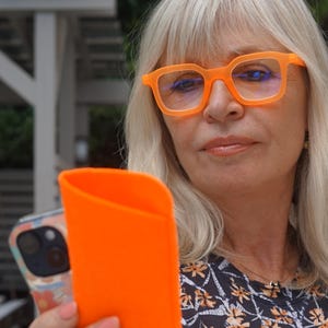 May include: A woman wearing bright orange eyeglasses with a matching orange case. The eyeglasses have a square frame. The woman is wearing a dark top with a floral pattern. Her hair is blonde.