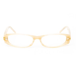 以下が含まれることがあります： 薄いベージュ色のフレームとクリアレンズの眼鏡。長方形のフレームには繊細な模様があり、テンプルには金色のディテールが施されています。眼鏡は白い背景に置かれています。