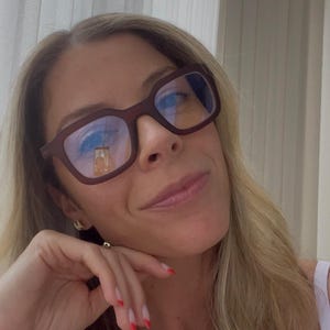 May include: A close-up shot of a person wearing brown-framed glasses with blue light lenses. The person has blonde hair and is smiling. The glasses reflect a blurry image. The person is wearing a white top and has red nail polish.