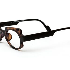 May include: A pair of eyeglasses with a distinctive design. The frames feature a tortoiseshell pattern, and the arms are solid black. The text "E-MODA ITALY DESIGN" is printed on the frame. The glasses are set against a white background.