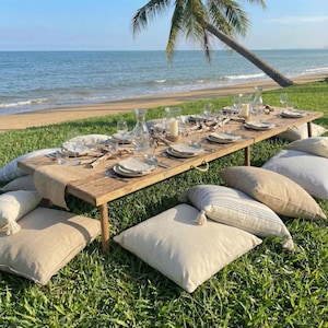 Könnte beinhalten: Ein niedriger hölzerner Picknicktisch auf einer Wiese am Strand mit einem Jutetischläufer, weißen Tellern, klaren Gläsern und Kerzen. Der Tisch ist von zahlreichen quadratischen Bodenkissen mit beigen und weißen Streifenmustern umgeben.
