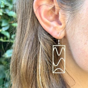 May include: Silver rectangular earrings with a wavy design inside. The earrings are hanging from a person's ear. The earrings are a simple, modern design.