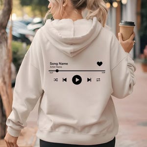 May include: A cream-coloured hoodie with a black music player graphic on the back. The graphic includes a heart, play, pause, and skip buttons. The text on the graphic reads "Song Name" and "Artist Name".