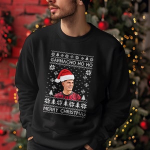 Könnte beinhalten: Schwarzes Sweatshirt mit weihnachtlichem Design. Das Design enthält den Text "GARNACHO HO HO" und "MERRY CHRISTMAS" in Weiß sowie ein Porträt einer Person mit Weihnachtsmütze. Ein festliches Kleidungsstück.