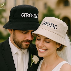 Peut inclure: Deux chapeaux de pêcheur, un noir avec le mot "GROOM" brodé en blanc et un beige avec "BRIDE" brodé en bleu marine. Le couple porte les chapeaux, célébrant leur mariage.
