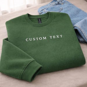 May include: A folded dark green sweatshirt with the text "CUSTOM TEXT" in white. Light blue denim jeans are next to the sweatshirt. The sweatshirt is on a neutral-colored surface.