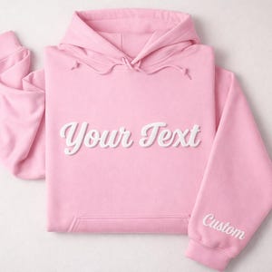 May include: A light pink hoodie with a drawstring hood and a kangaroo pocket. The words "Your Text" are displayed in a white, cursive font across the chest. The right sleeve has the word "Custom" in white.